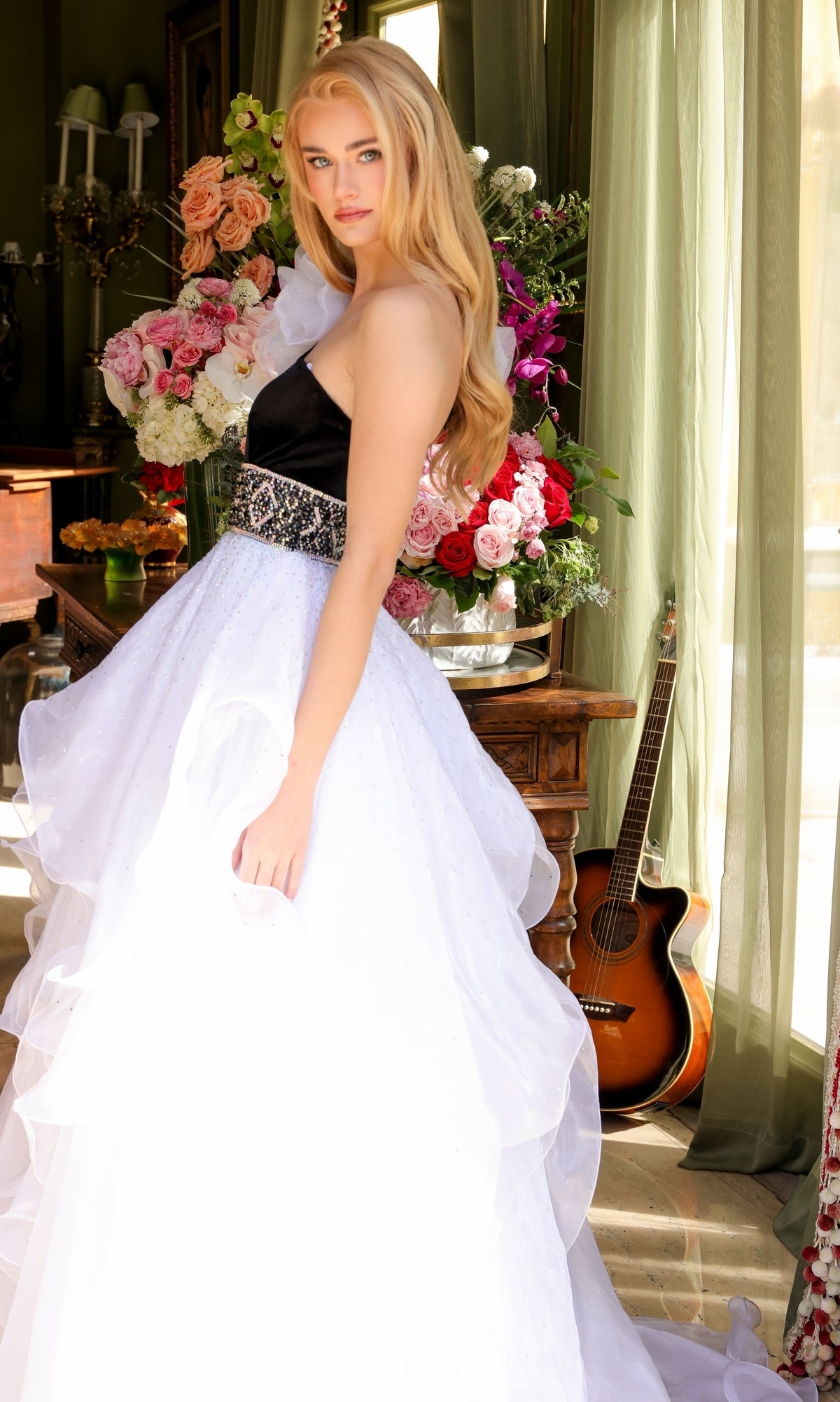 A woman wears the Ava Presley Long Prom Dress 39557, a strapless black and white gown with a layered skirt, standing indoors near large flower arrangements and a guitar by a window with green curtains.